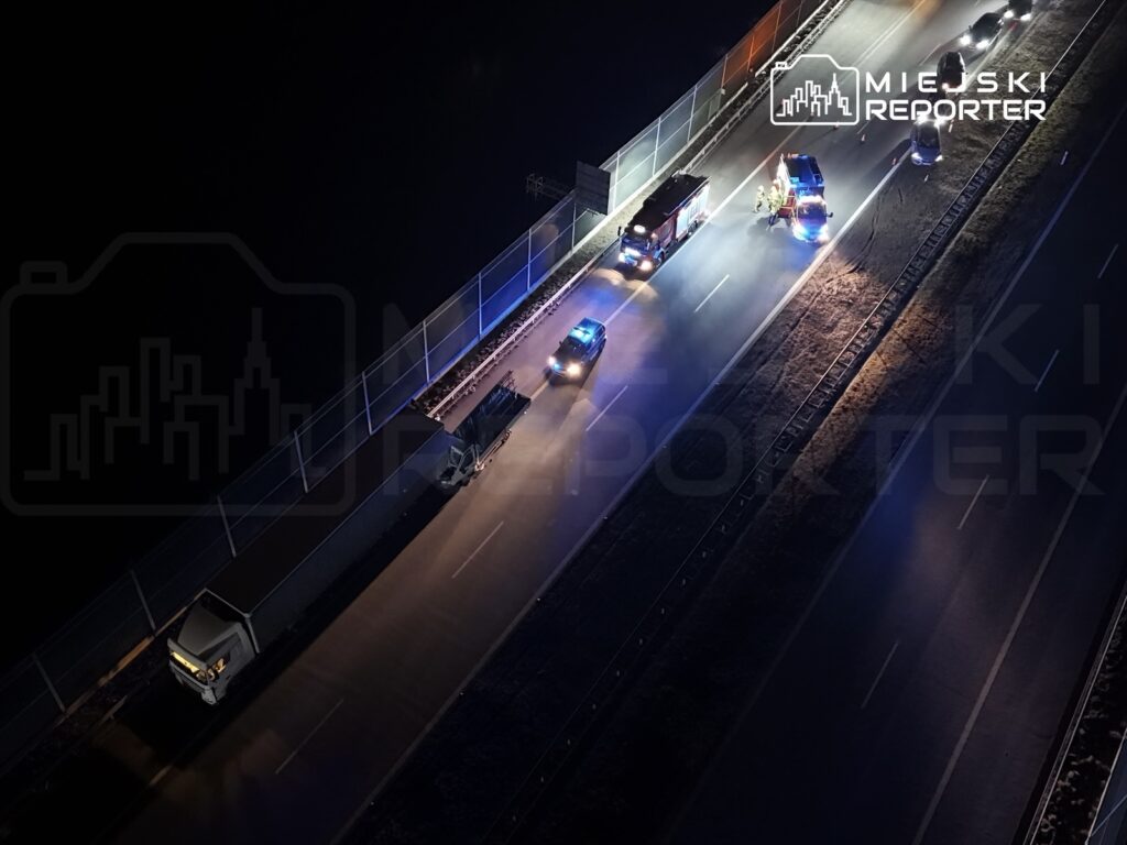 Na drodze widoczne są samochody policyjne i strażackie, które zatrzymały ruch przy unieruchomionym pojeździe ciężarowym.