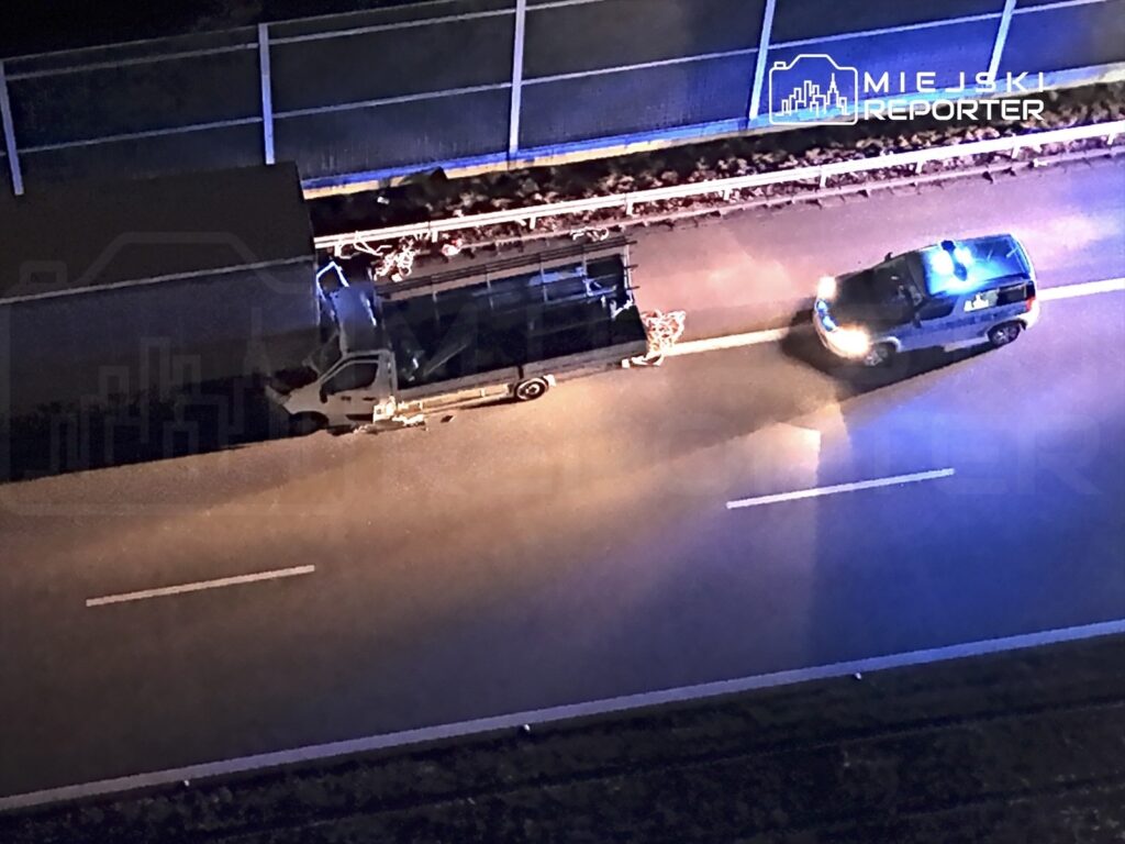 Biały samochód dostawczy zatrzymany na drodze, obok niego radiowóz policyjny z włączonymi sygnałami świetlnymi.