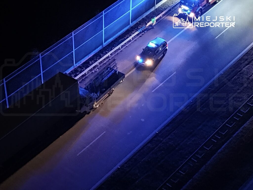 Na drodze stoi ciężarówka, obok niej zaparkowane są radiowozy policyjne z włączonymi sygnałami świetlnymi.