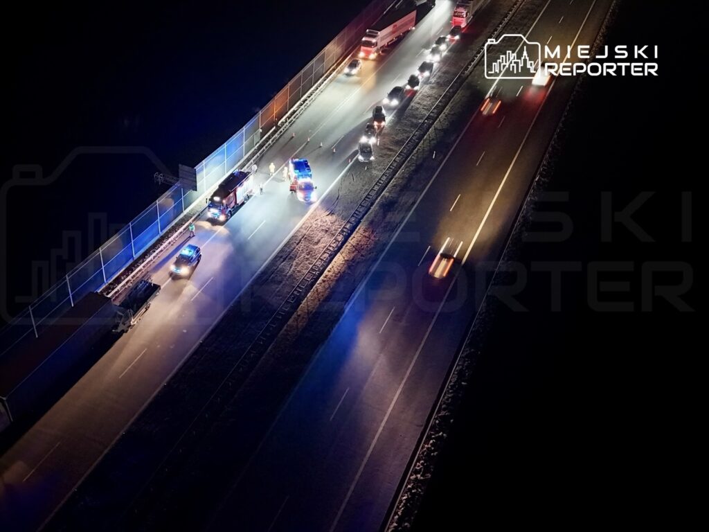 Na drodze nocą, pojazdy służb ratunkowych zatrzymane obok bariery, oświetlone niebieskimi i czerwonymi światłami.