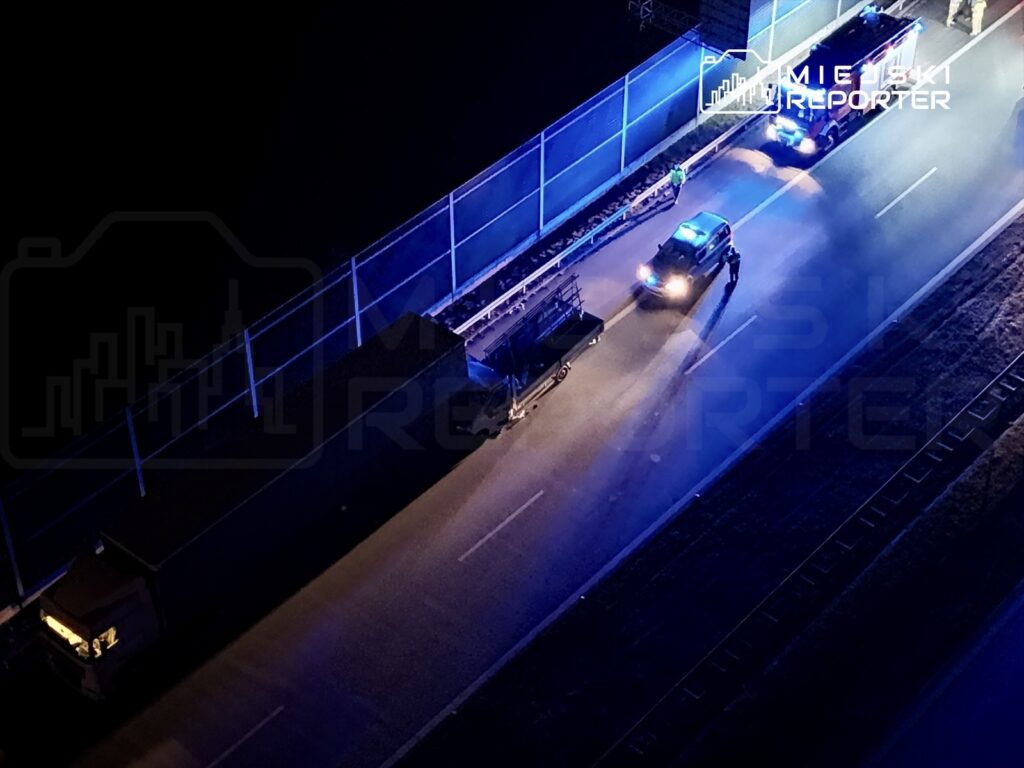 Policyjny radiowóz z włączonymi sygnałami świetlnymi zatrzymuje pojazd na drodze w nocy, obok płotu.