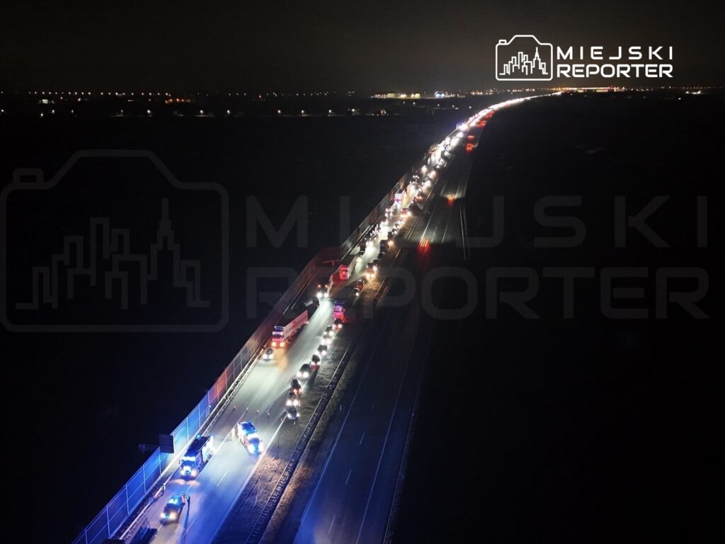 Ruch samochodowy w nocy na drodze, z policyjnymi pojazdami włączonymi sygnałami świetlnymi.