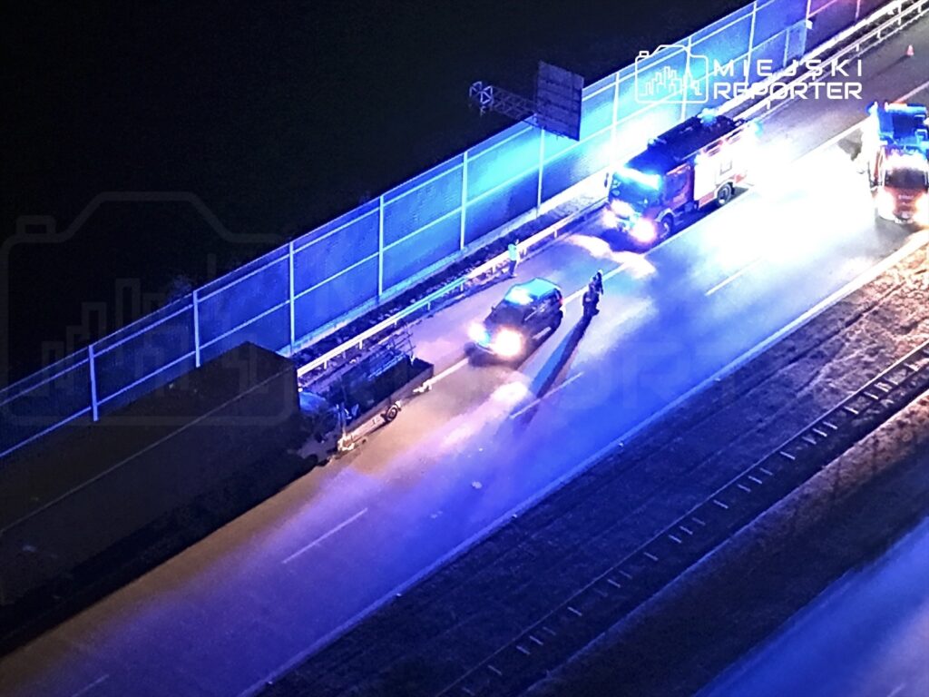 Pojazdy służb ratunkowych oraz policyjnych zatrzymane przy oświetlonej drodze, obok stoi ciężarówka.