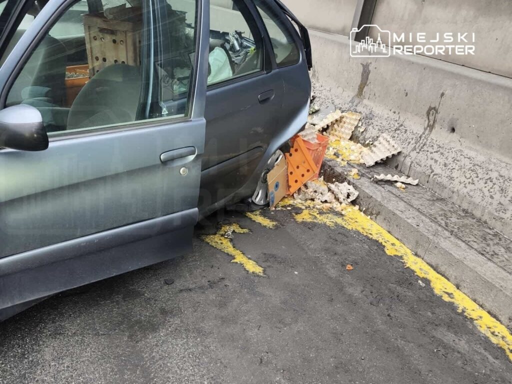 Uszkodzony samochód zaparkowany przy ścianie, z porzuconymi odpadami i kartonami leżącymi obok.