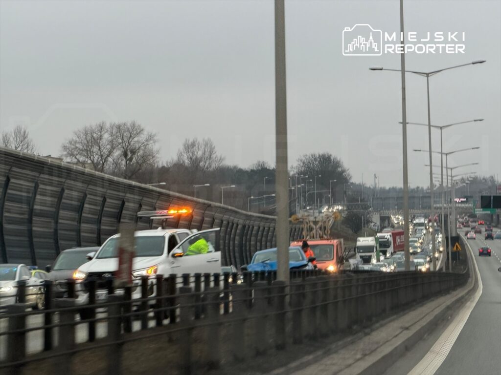 Na drodze stoi samochód z włączonymi światłami awaryjnymi, obok pracownik w żółtej kamizelce. W tle widać zator drogowy.