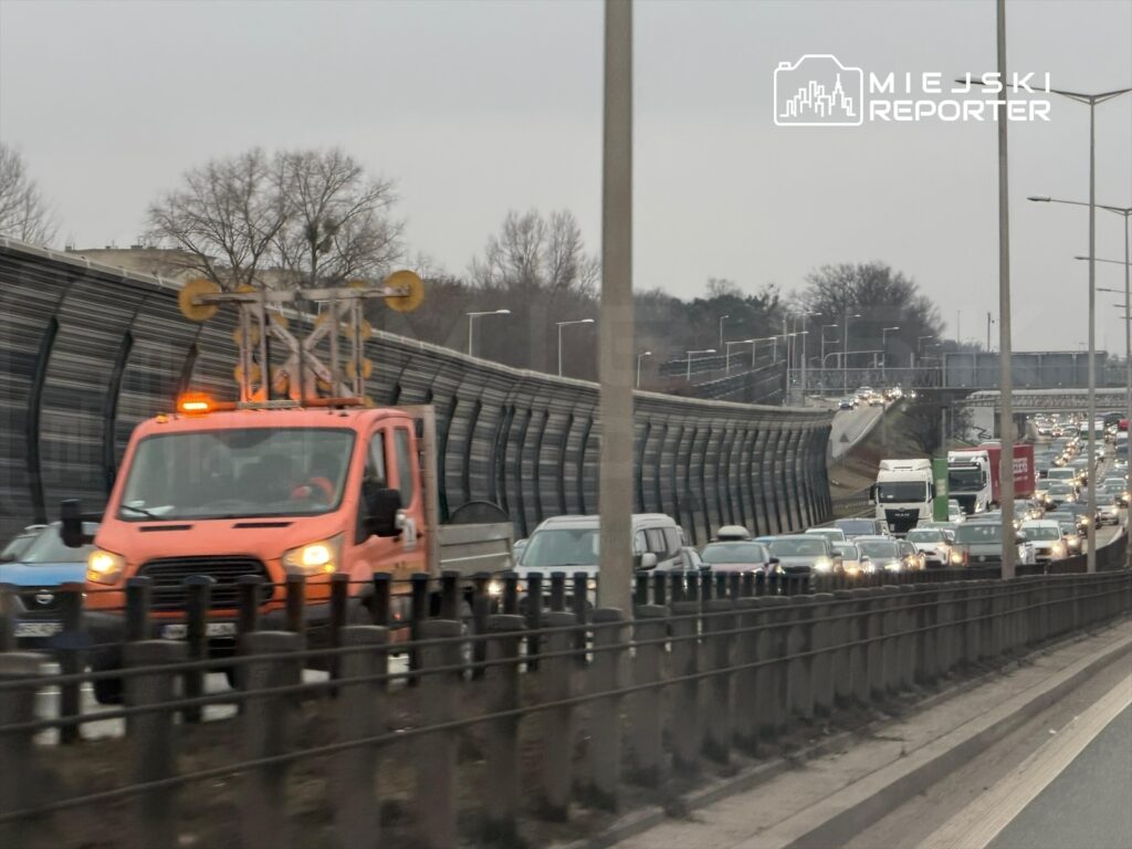 Pomarańczowy pojazd serwisowy z sygnalizacją świetlną przemieszcza się wzdłuż zakorkowanej drogi, obok ogrodzenia dźwiękochłonnego.
