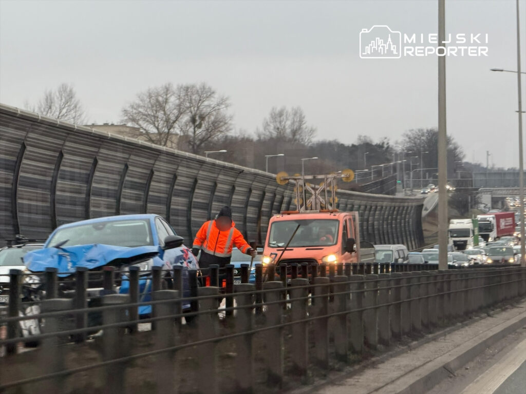 Mężczyzna w pomarańczowej kamizelce pracuje przy uszkodzonym niebieskim samochodzie na ruchliwej drodze.
