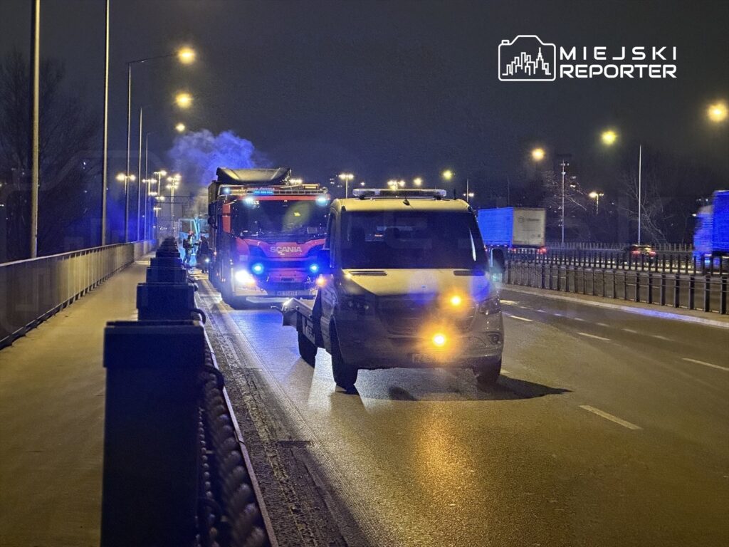 Czerwony ciężarowy pojazd z włączonymi sygnałami świetlnymi zatrzymany na drodze w nocy, obok stoi biały furgon.
