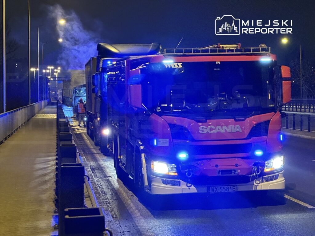 Czerwony wóz strażacki z włączonymi sygnalizacjami świetlnymi stoi na drodze, obok unieruchomionego pojazdu ciężarowego.