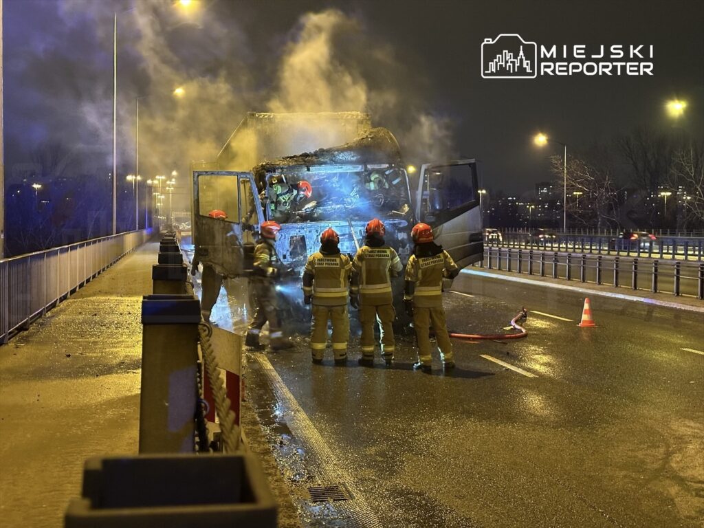 Strażacy w hełmach gaszą pożar w uszkodzonym pojeździe na moście w nocy, otoczeni dymem i światłami.