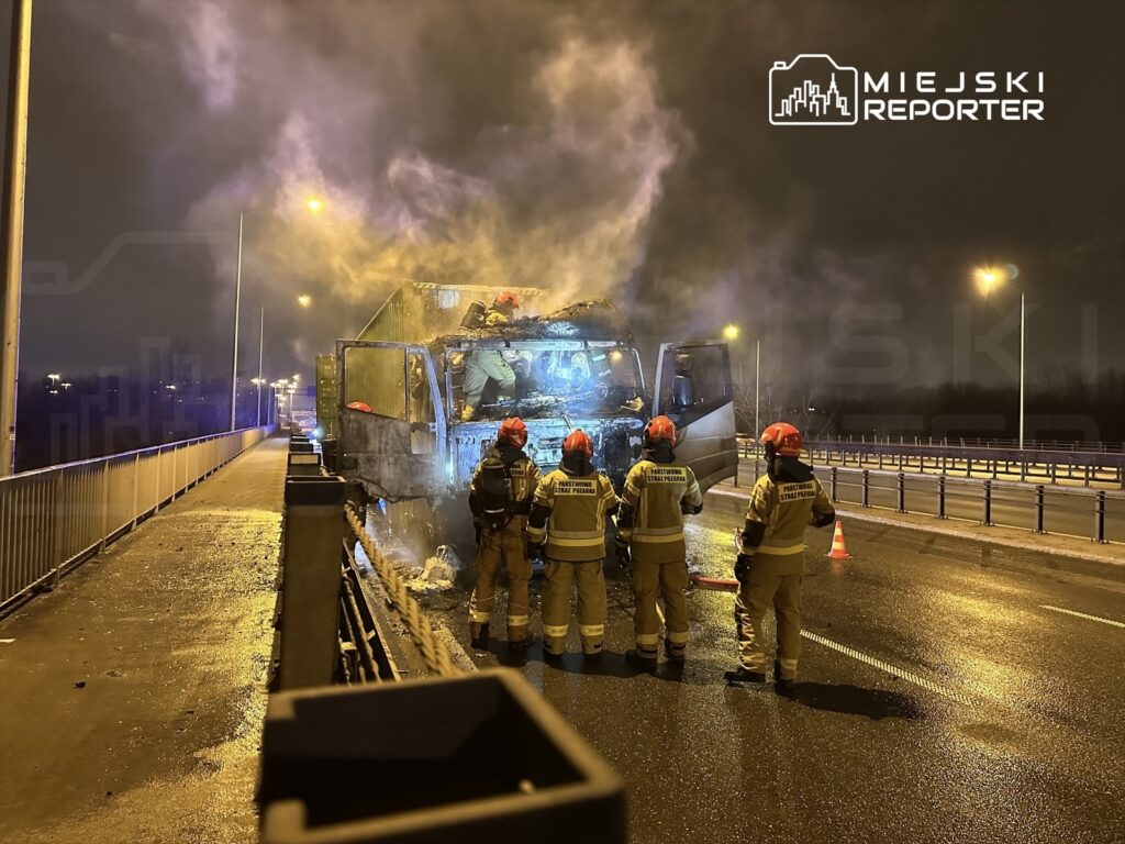 Strażacy w hełmach gaszą płonący pojazd na moście w nocy, wokół unoszą się kłęby dymu.