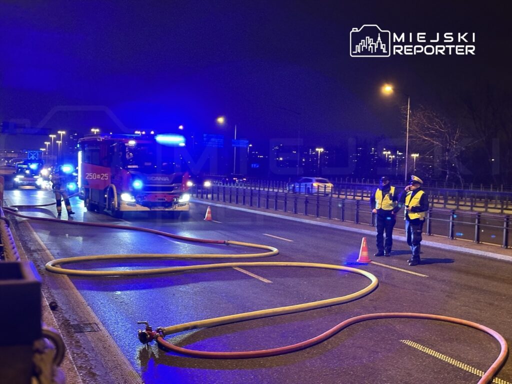 Strażacy przy wozie strażackim rozkładają węże na jezdni, podczas nocnej akcji ratunkowej w miejskim otoczeniu.
