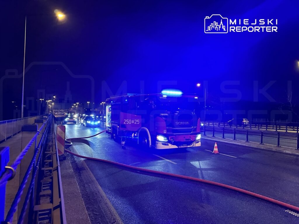 W nocy straż pożarna z włączonymi sygnałami świetlnymi rozstawia węże na drodze, obok stoją samochody osobowe.