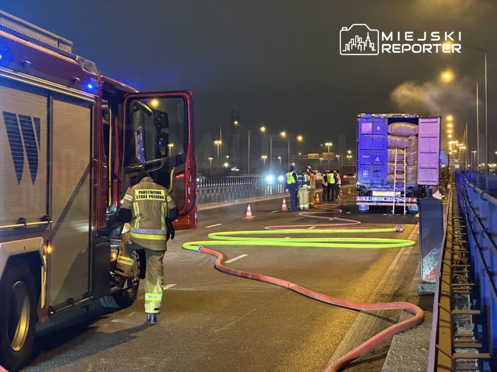 Strażacy w mundurach prowadzą działania ratunkowe przy ciężarówce na moście, w tle widoczne światła policyjne.