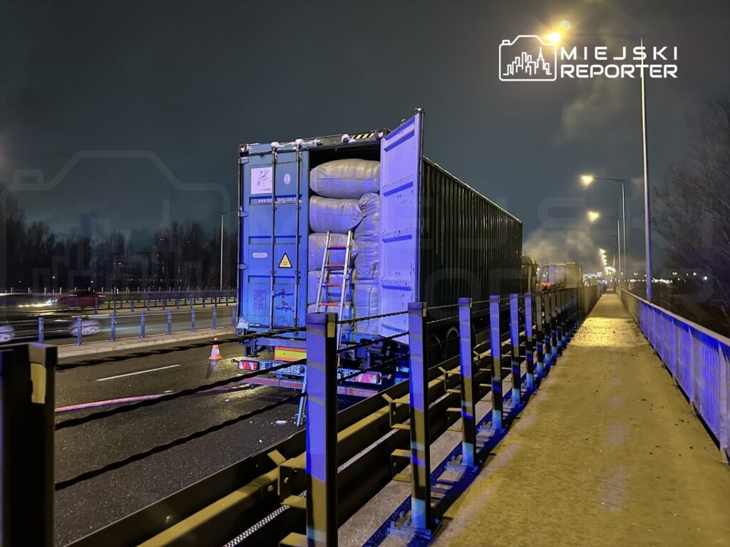 Kontener na ciężarówce stoi na pasie ruchu, obok widoczne są bariery ochronne i znaki ostrzegawcze w nocy.