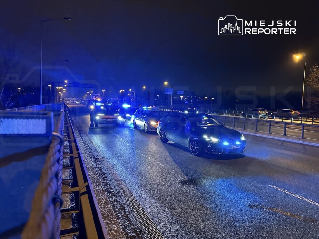 Na drodze nocą stoją samochody policyjne z włączonymi sygnałami świetlnymi, a funkcjonariusz rozmawia z kierowcą.