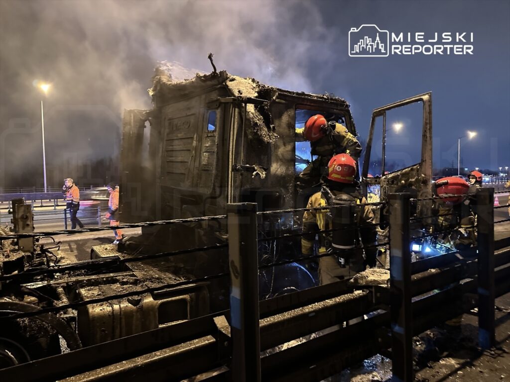 Strażacy gaszą pożar spalonej ciężarówki na autostradzie w nocy, w otoczeniu dymu i płomieni.