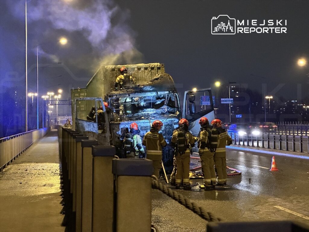 Strażacy w hełmach gaszą ogień w spalonej ciężarówce na moście w nocy, otoczeni dymem i niebieskimi światłami.