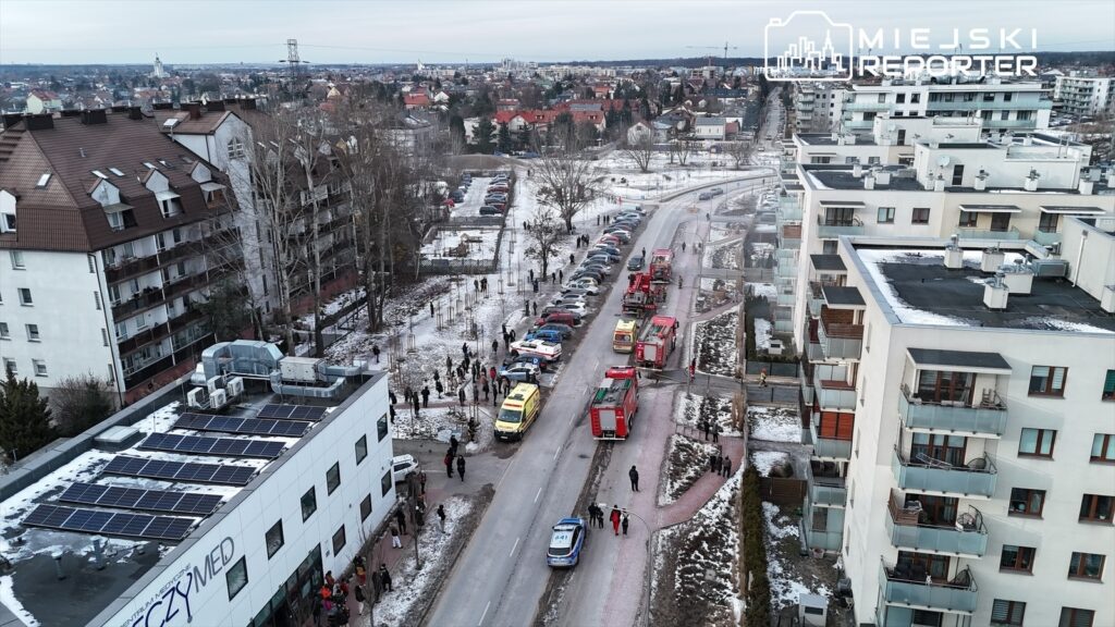 Zespół służb ratunkowych, w tym karetki i wozy strażackie, interweniuje na ulicy, gdzie zgromadził się tłum ludzi. W tle widać nowoczesne budynki mieszkalne oraz pokryty śniegiem teren.