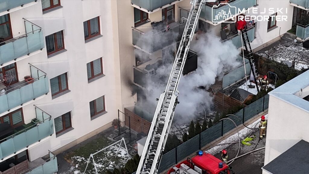 Strażacy gaszą pożar z wykorzystaniem drabiny, w tle widoczne dymiące mieszkanie w budynku wielorodzinnym.