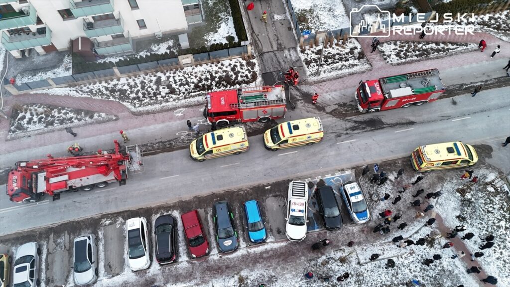 Strażacy z wozem gaśniczym oraz karetki pogotowia na ulicy, obok parkujących samochodów, w otoczeniu gapiów.