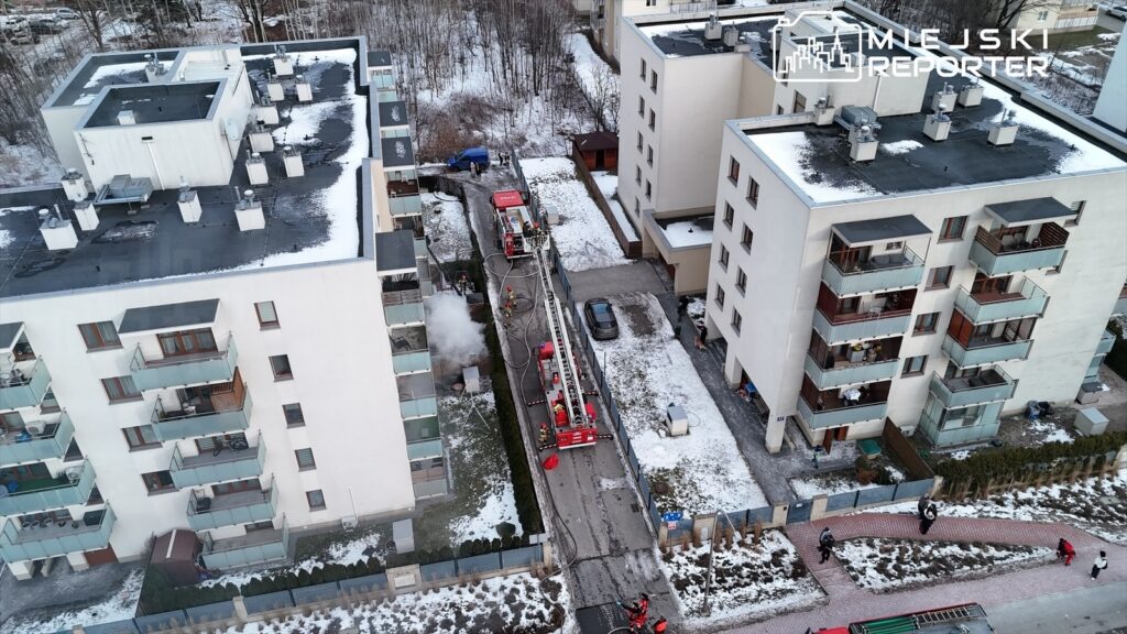 Strażacy z wozem gaśniczym pracują przy budynku mieszkalnym, gdzie widać dym wydobywający się z terenu.