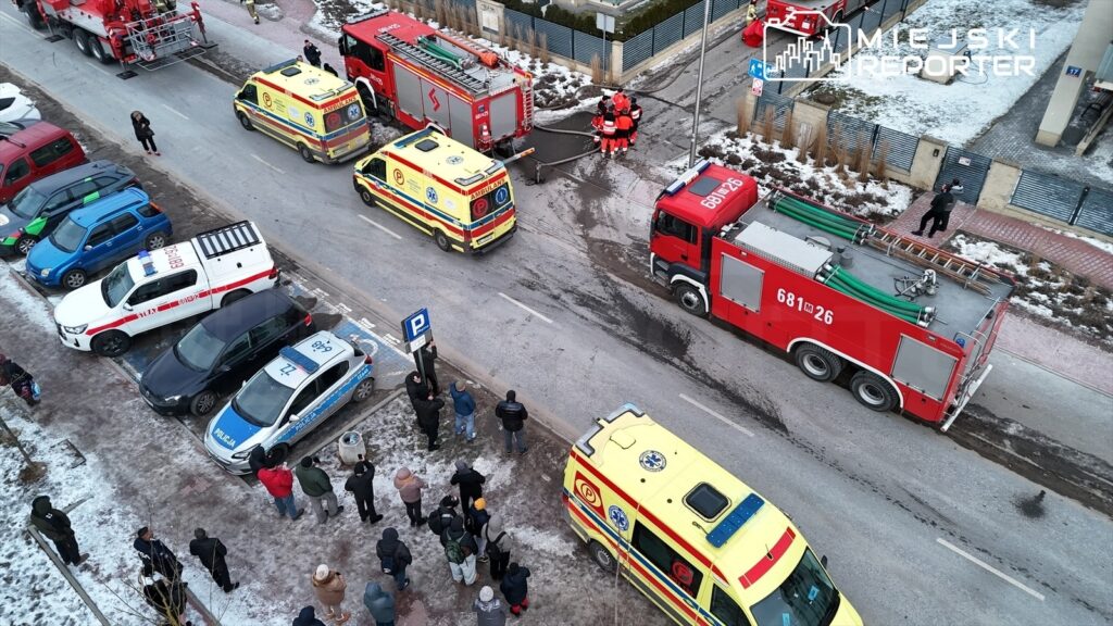 Grupa ratowników w odblaskowych strojach omawia działania obok pojazdów straży pożarnej i karetek na zaśnieżonej ulicy.