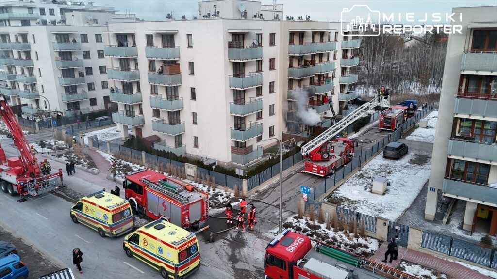 Strażacy używają drabiny, aby ugasić dym wydobywający się z balkonu w bloku mieszkalnym, obok parkują karetki.