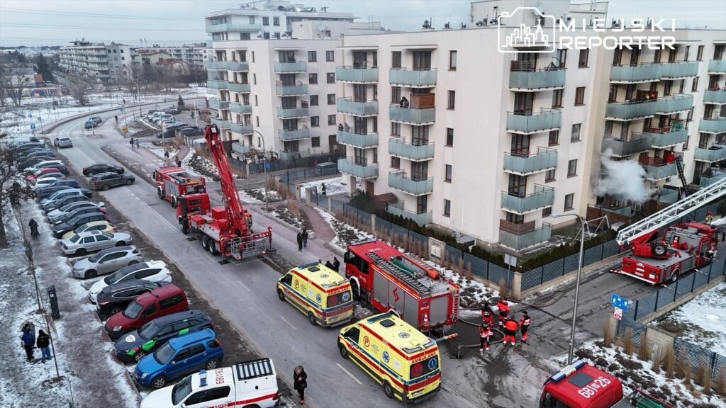 Zespół ratunkowy w czerwonych mundurach pracuje przy bloku mieszkalnym, gdzie wydobywa się dym.