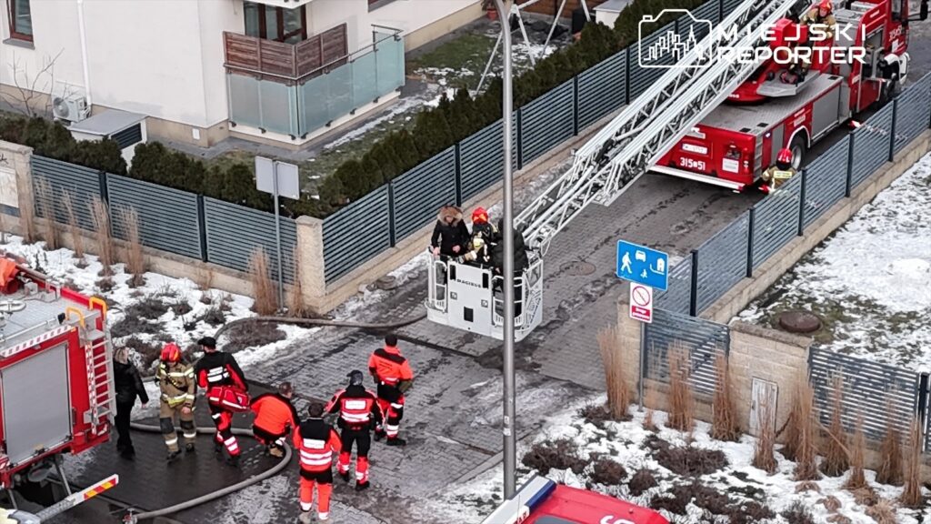 Grupa strażaków w odzieży ochronnej prowadzi akcję ratunkową, podczas gdy jeden z nich wchodzi na podnośnik.