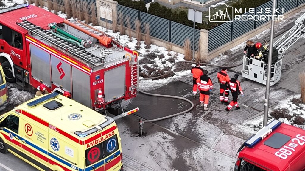 Grupa ratowników w pomarańczowych strojach omawia sytuację obok wozu strażackiego i karetki na zaśnieżonej ulicy.