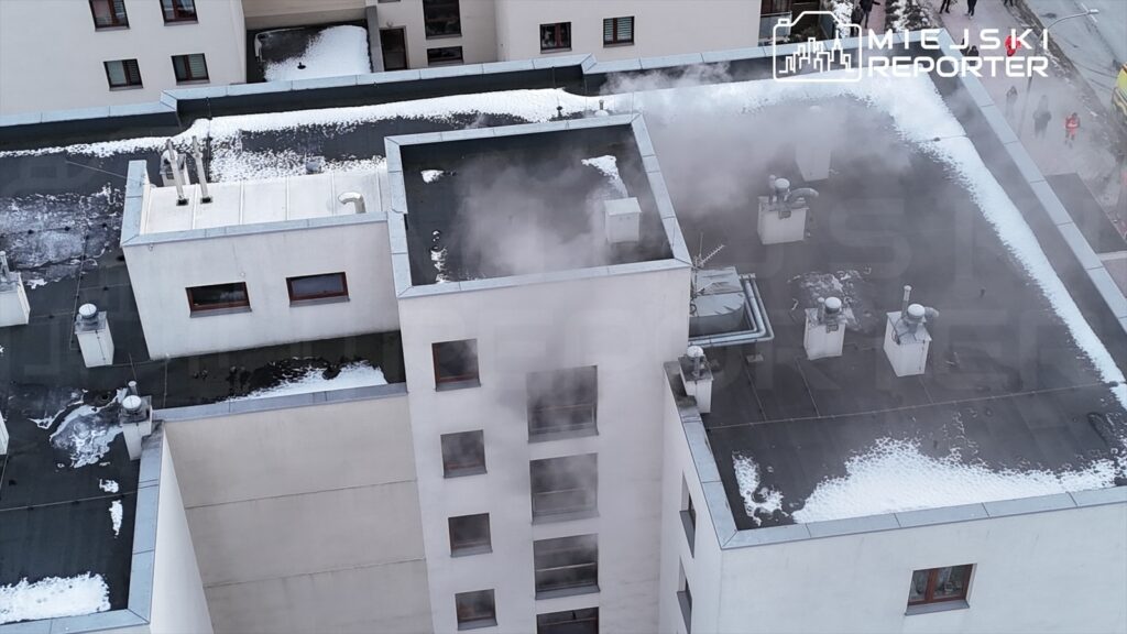 Na dachu budynku unosi się gęsty dym, a wokół widoczne są resztki śniegu.