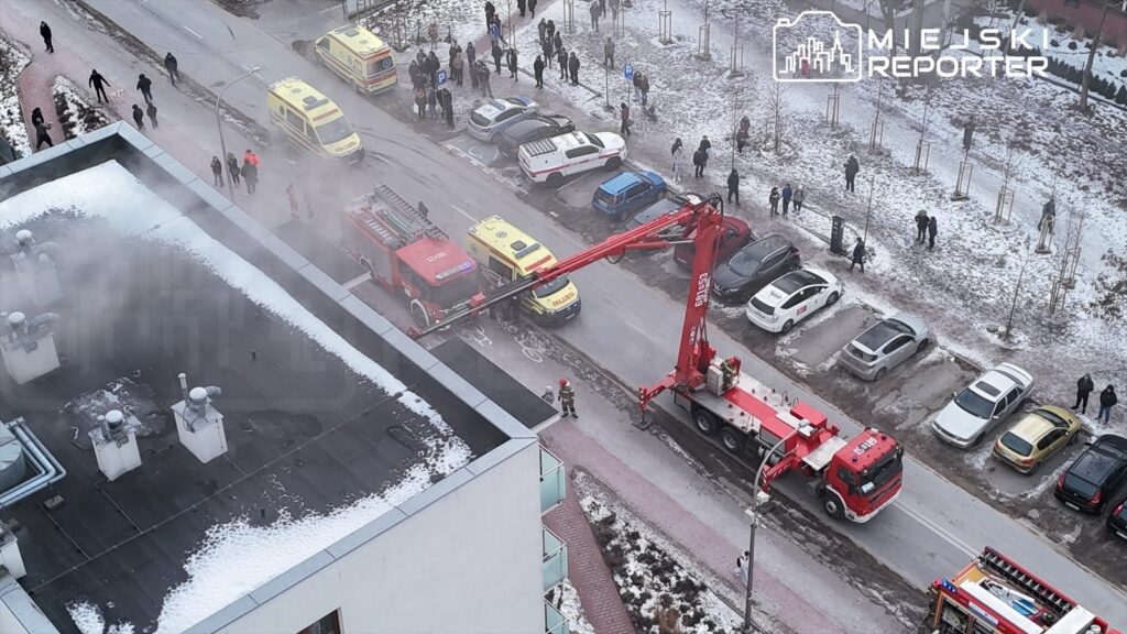 Strażacy z wozem gaśniczym używają podnośnika do gaszenia pożaru na dachu budynku, w otoczeniu ludzi i pojazdów.