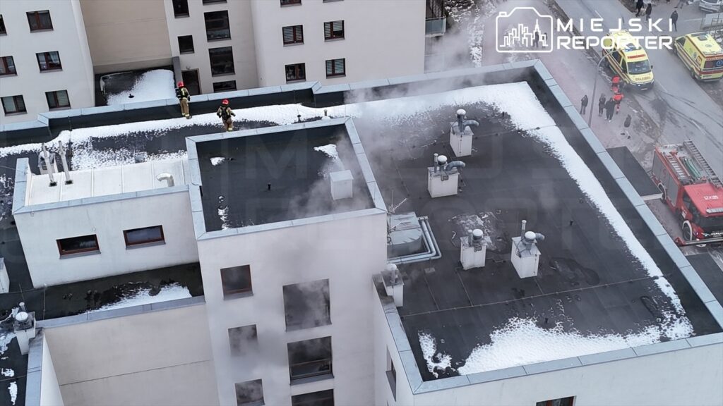 Dwaj strażacy w hełmach gaszą dymiący dach nowoczesnego budynku mieszkalnego w mieście. W tle widać wozu strażackie.