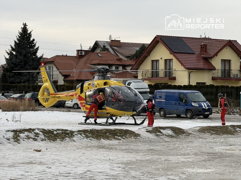 Ratownicy w czerwonych strojach przygotowują się do startu helikoptera w zimowym otoczeniu, obok domów.