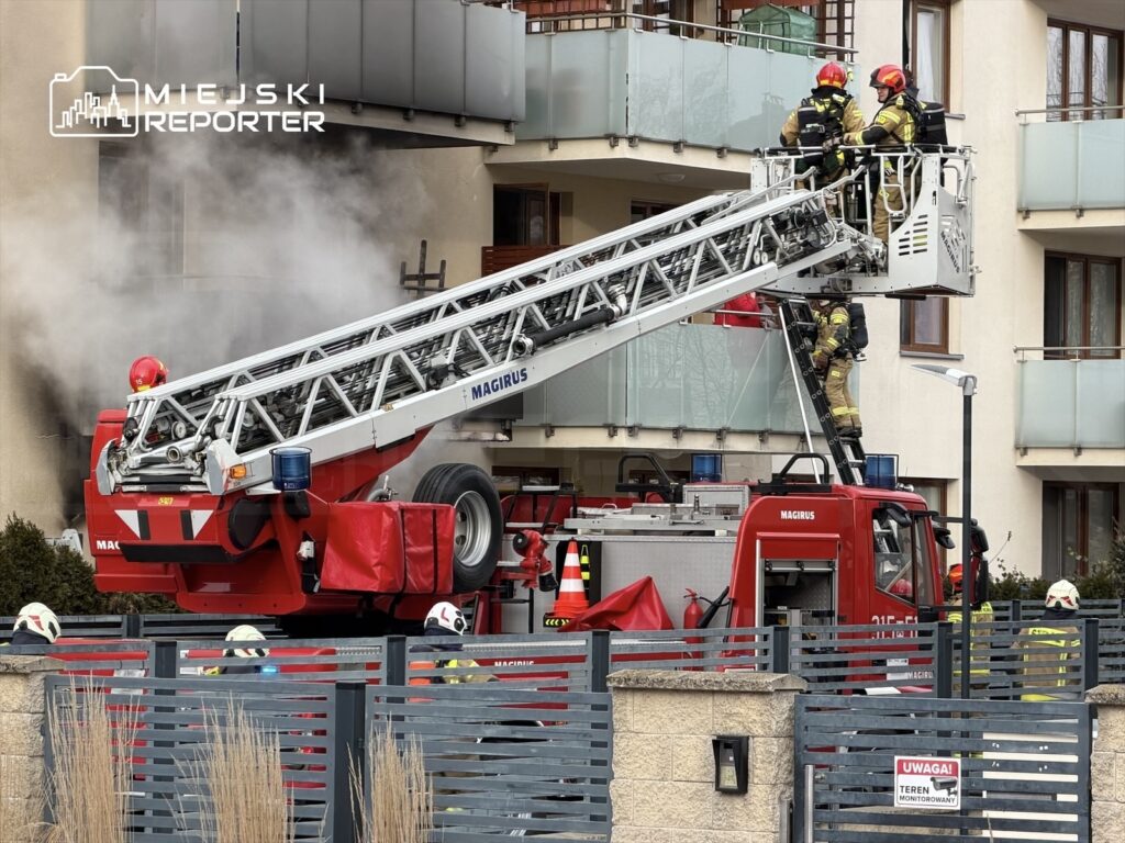 Strażacy na wysięgniku w akcji gaszenia pożaru w budynku mieszkalnym, otoczeni przez pojazdy ratunkowe.