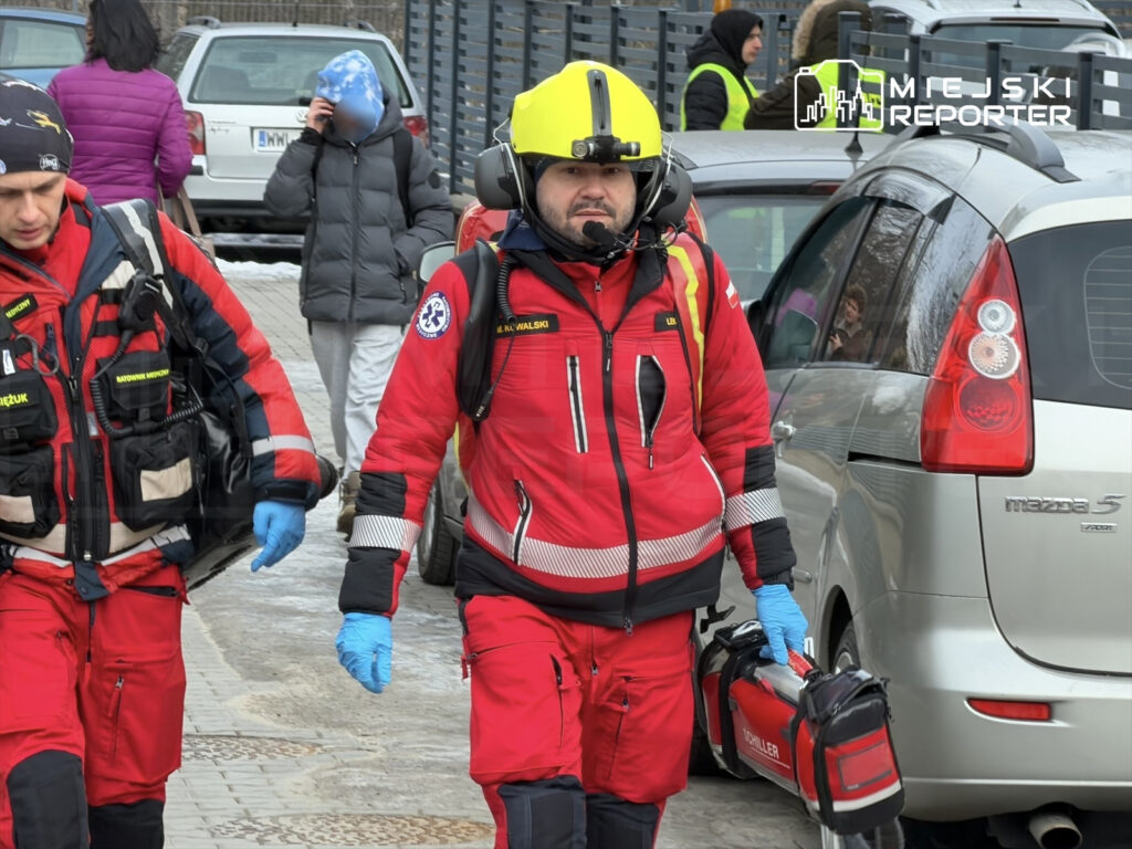 Ratownicy medyczni w czerwonych uniformach niosą sprzęt medyczny na chodniku, w tle samochody i przechodnie.