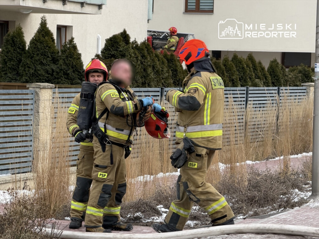 Dwaj strażacy w mundurach rozmawiają na chodniku, trzymając sprzęt ratunkowy, w tle widać budynek mieszkalny.
