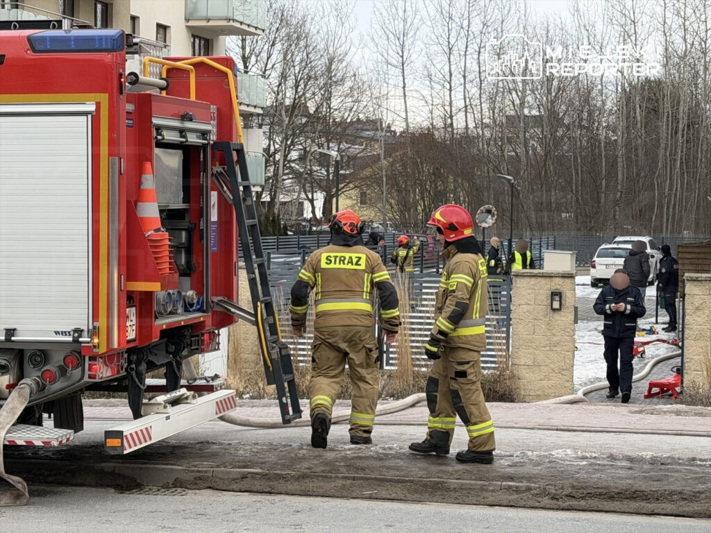 Strażacy w mundurach i hełmach przygotowują się do akcji ratunkowej obok wozu strażackiego na osiedlu.