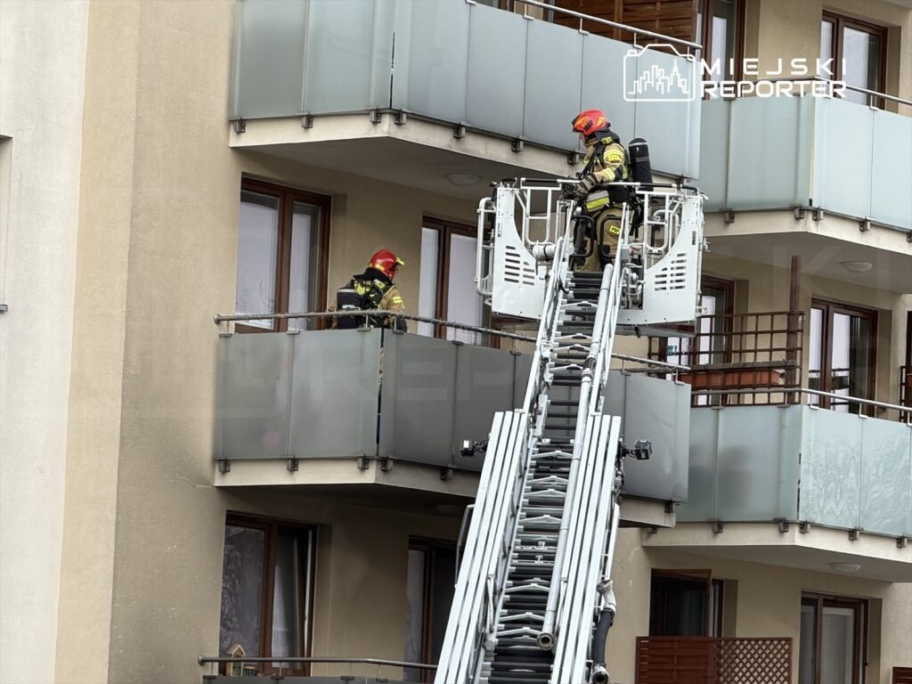 Strażacy na podnośniku wykonują prace ratunkowe na balkonie nowoczesnego budynku mieszkalnego.
