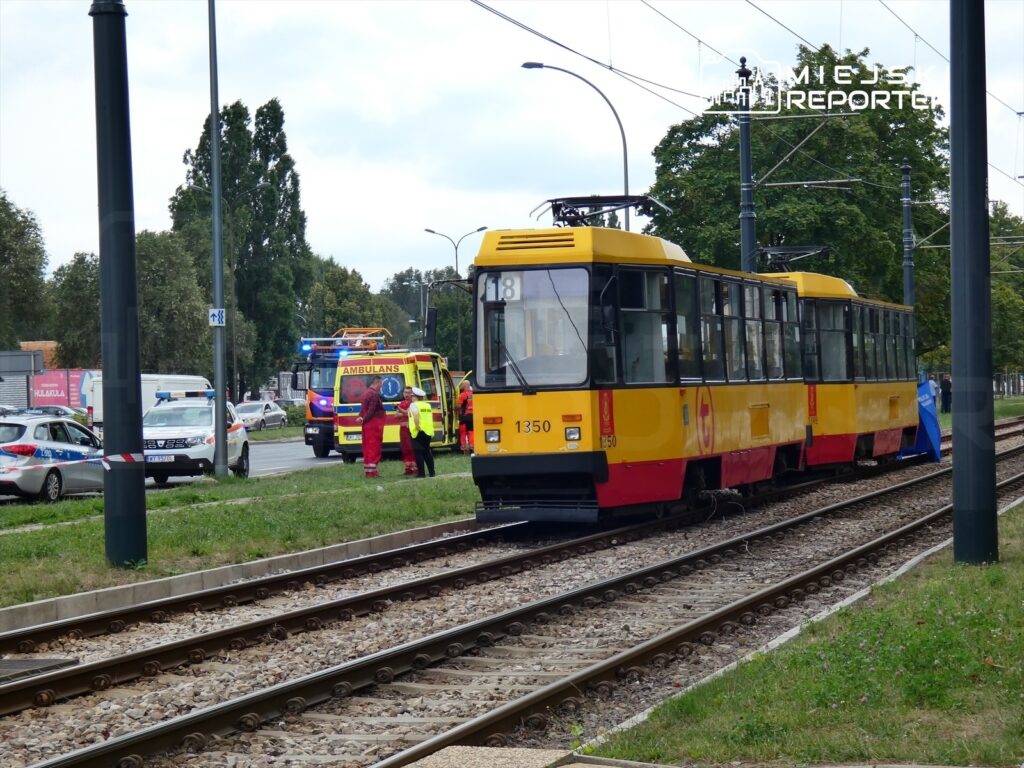 Tramwaj zatrzymany na torowisku, obok grupy ratowników w odzieży ochronnej oraz pojazdów służb ratunkowych.