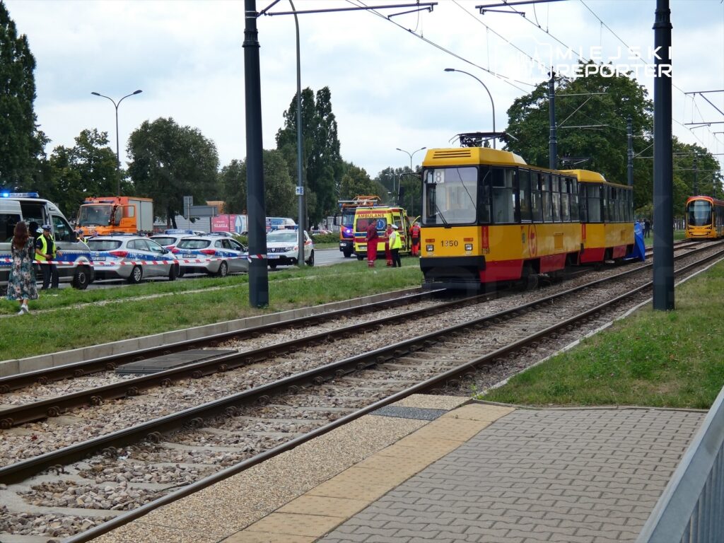 Tramwaj zatrzymany na torowisku obok policyjnych radiowozów oraz pojazdów służb ratunkowych na miejskiej ulicy.