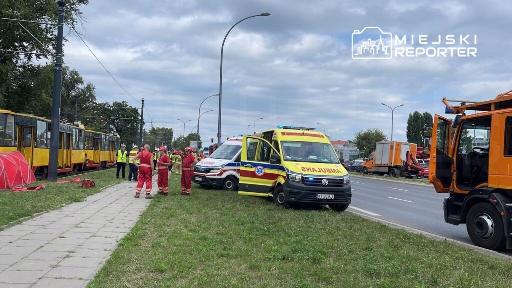 Grupa ratowników w czerwonych strojach omawia sytuację obok dwóch karetek pogotowia na trawniku przy torach tramwajowych.
