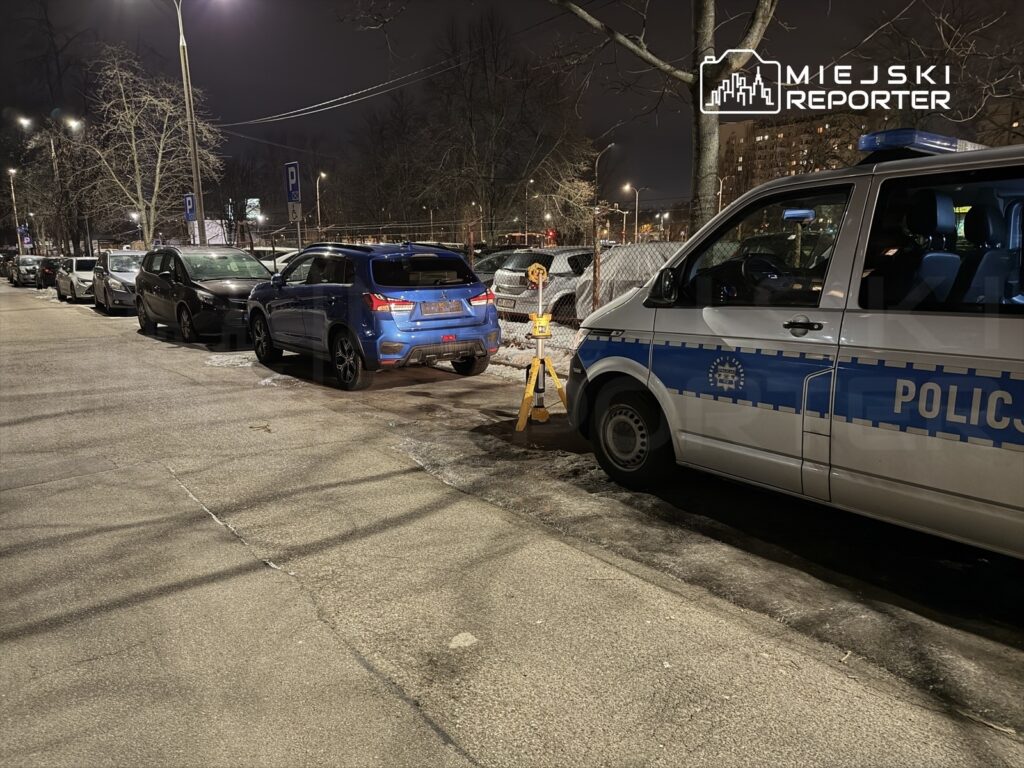 Policyjny furgon zaparkowany obok nieprawidłowo stojącego niebieskiego SUV-a na oświetlonej ulicy nocą.
