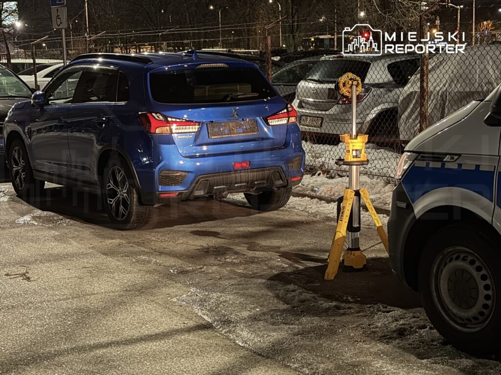 Niebieski samochód osobowy zaparkowany obok słupa z oświetleniem, w tle widoczny inny pojazd i ogrodzenie.