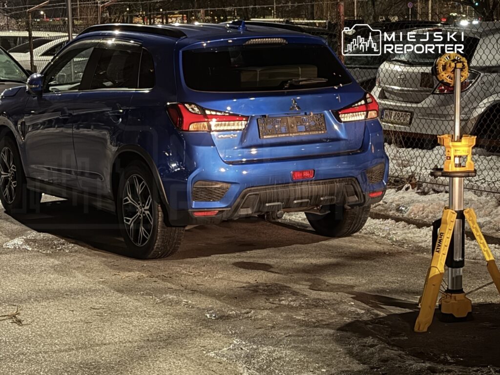 Niebieski SUV zaparkowany na oświetlonym parkingu obok żółtego statywu pomiarowego.