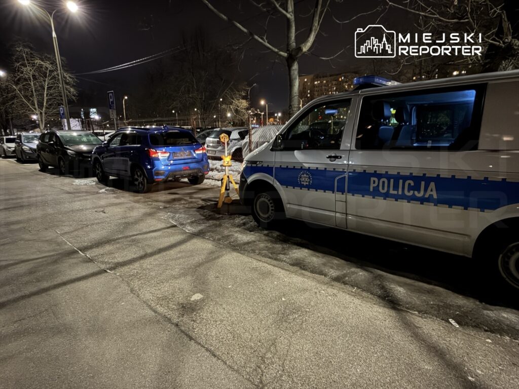 Policyjny radiowóz zaparkowany na oświetlonej ulicy, obok stoją dwa zaparkowane pojazdy. W tle widoczne budynki.
