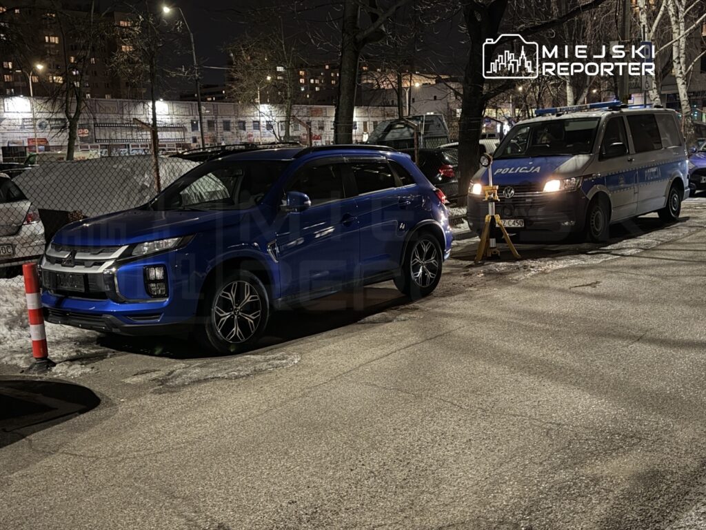 Niebieski SUV zaparkowany obok policyjnego vana na oświetlonej ulicy w nocy, w tle widać bloki mieszkalne.