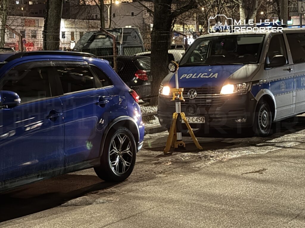 Nieoznakowany niebieski samochód osobowy stoi na ulicy obok radiowozu policyjnego z włączonymi światłami.