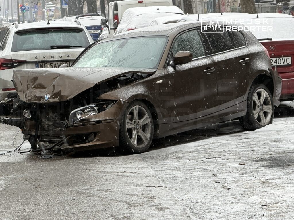 Uszkodzony brązowy samochód osobowy zaparkowany na zaśnieżonej ulicy, z widocznymi uszkodzeniami przodu.